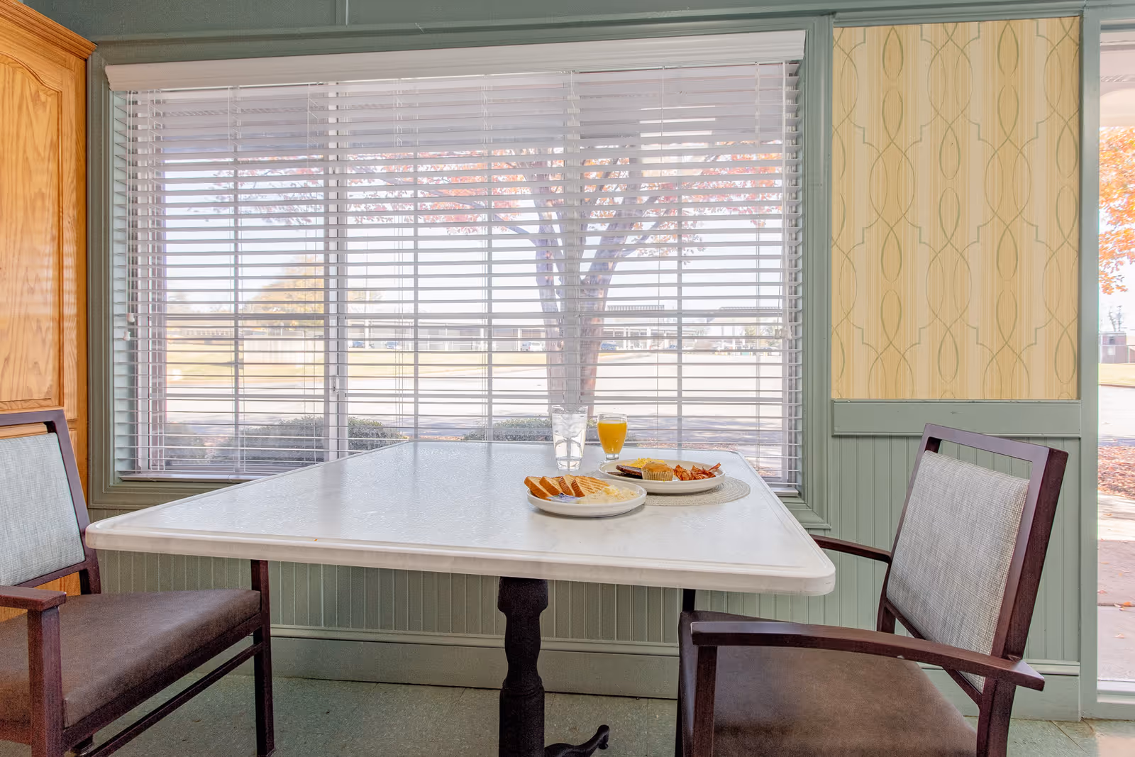A small dining area with a white table and two chairs with wooden frames and light gray cushions. On the table, there are two plates with breakfast food including toast, eggs, bacon, and a glass of orange juice and water. Behind the table is a large window with white blinds, showing an outdoor view with a tree and a building in the distance. The walls are painted green with a patterned wallpaper panel on the right side.