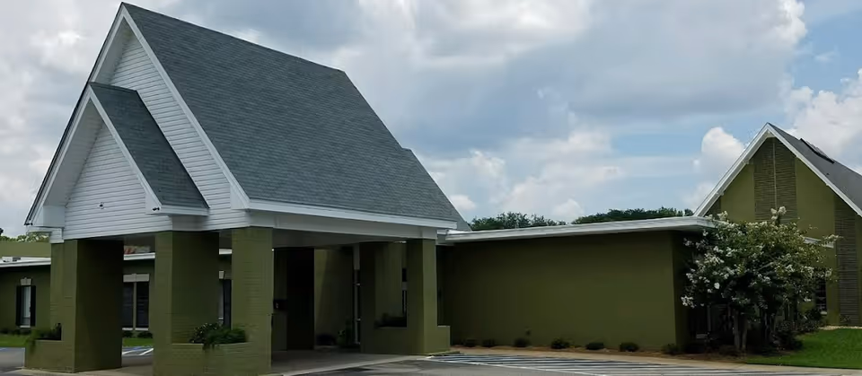 Front exterior of a green single-story building with a covered entrance/porte-cochere, steep gabled roof, and parking area.