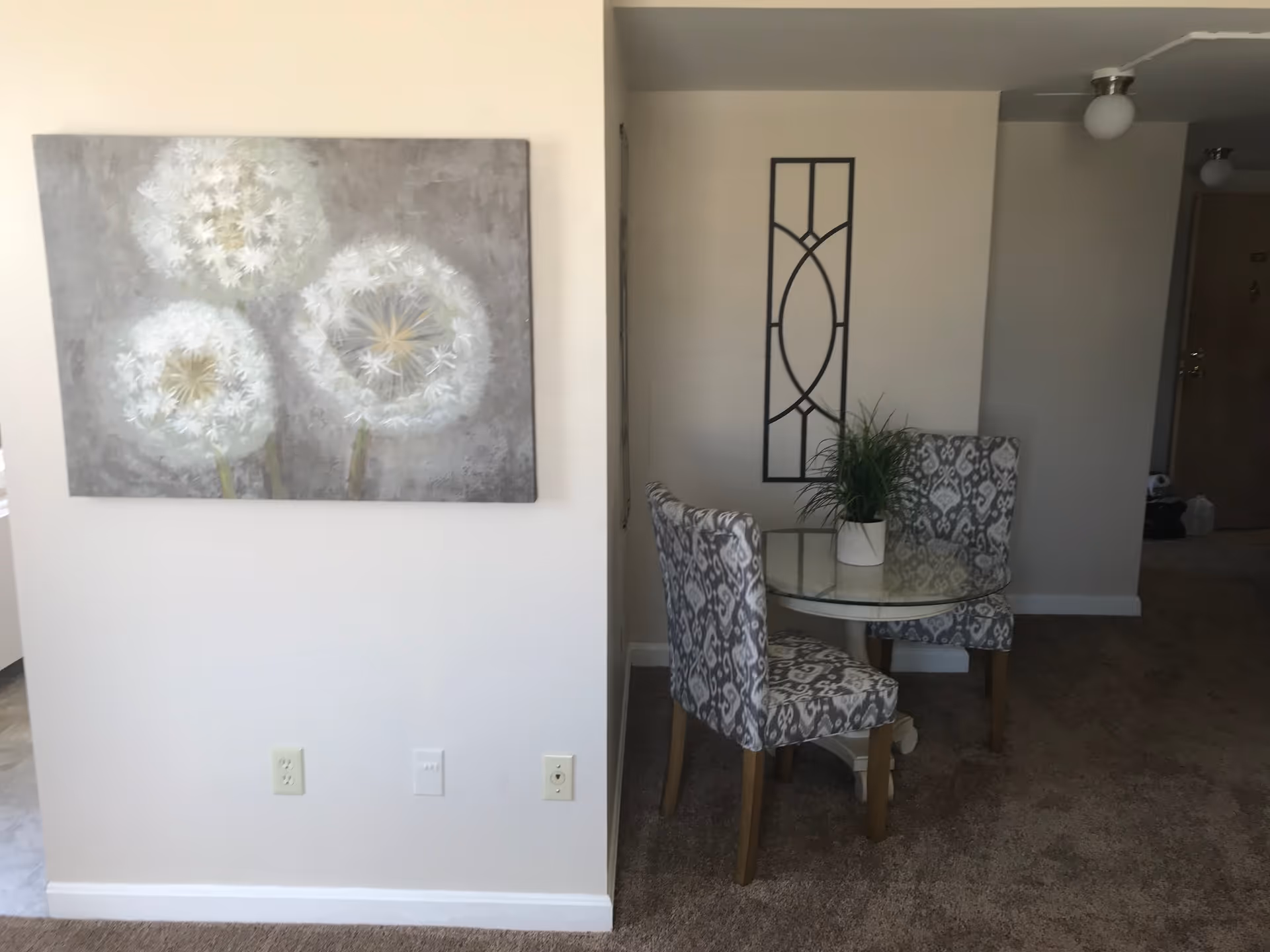 Interior view of a living space with a small round glass table and two patterned chairs. A potted plant is placed on the table. On the left wall, there is a large painting of three white dandelions. The floor is carpeted, and the walls are painted light beige. A decorative black metal wall art piece is hung on the wall behind the table.
