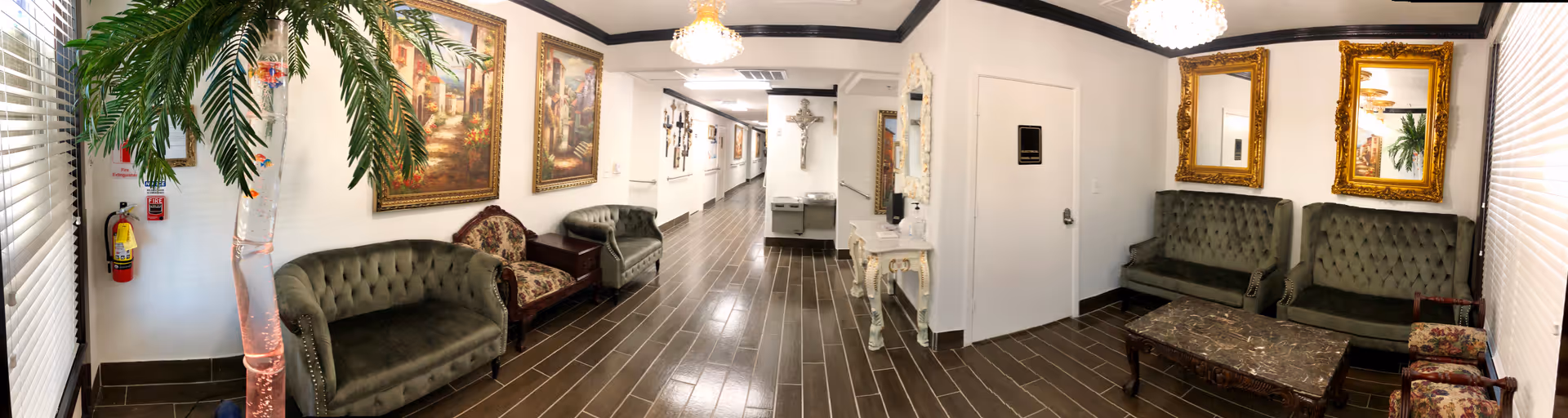 A long hallway in an assisted living facility with dark wood flooring and white walls. The hallway is furnished with multiple vintage-style green and patterned armchairs and sofas along the walls. There are several framed paintings and two large ornate gold-framed mirrors on the walls. A small white table with decorative items is placed near a door, and a large cross hangs on the wall at the end of the hallway. A tall artificial palm tree is positioned near a window with blinds on the left side.