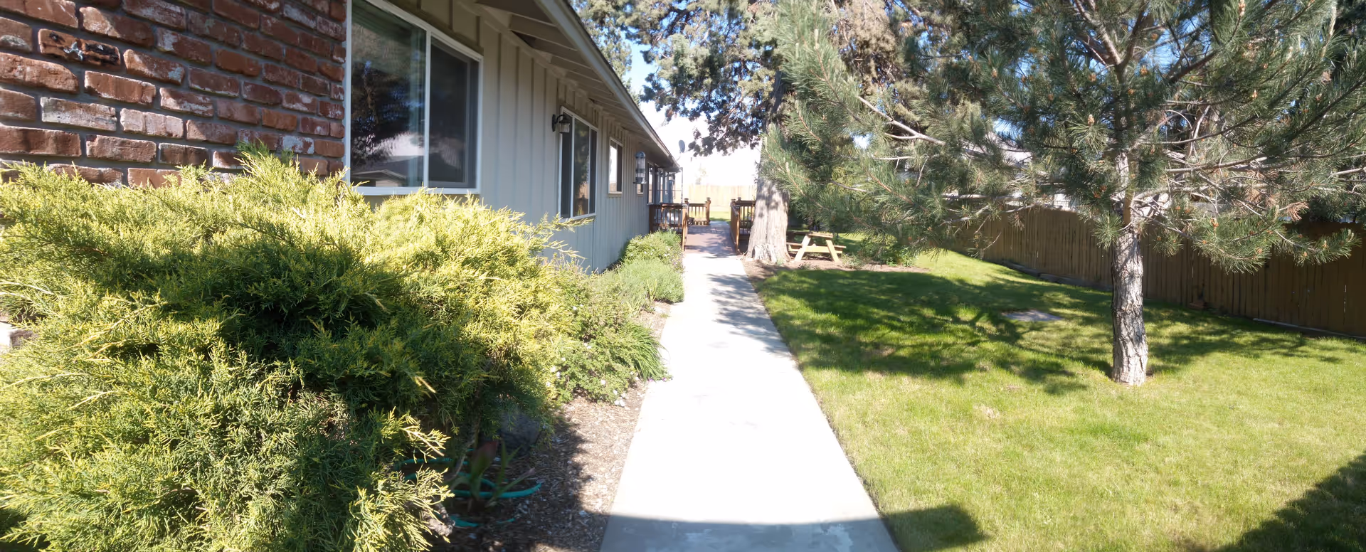 A sunny outdoor pathway alongside a building with brick and siding exterior walls. The path is bordered by green bushes on one side and a grassy area with trees and a wooden picnic table on the other side. A wooden fence encloses the yard.