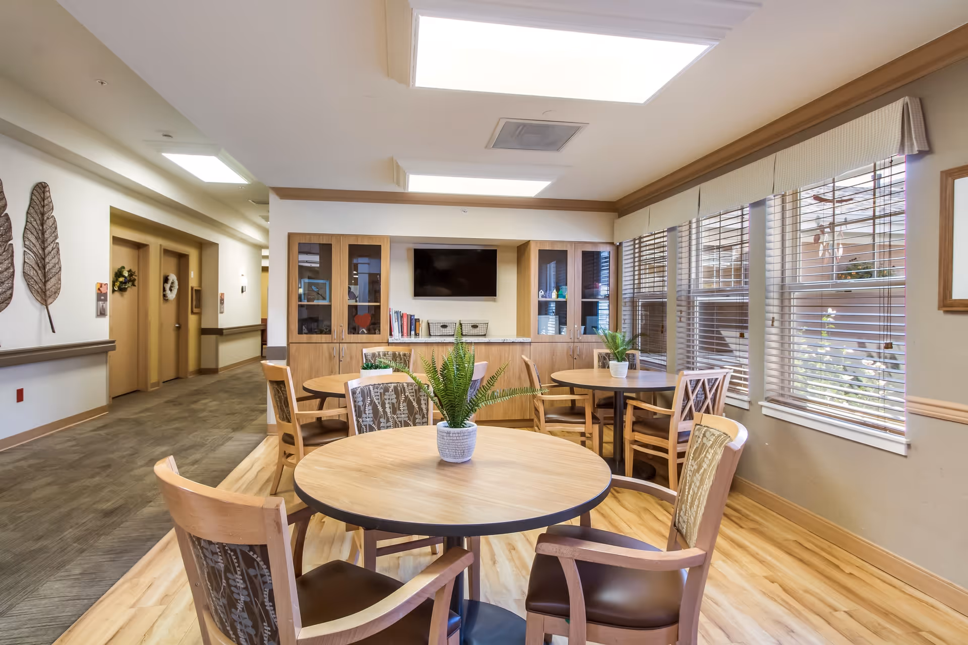 A bright and clean common area with round wooden tables and chairs arranged around them. Each table has a small potted plant. Large windows with blinds allow natural light to fill the room. In the background, there is a wooden cabinet with glass doors, books, and a flat-screen TV mounted above it. The floor is a combination of wood and carpet, and the walls are decorated with leaf-shaped wall art and framed pictures.