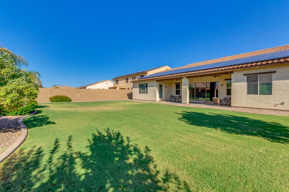 A spacious backyard with well-maintained green grass, bordered by a curved stone edge with bushes and trees on the left side. The backyard is adjacent to a single-story house with a covered patio area furnished with outdoor chairs and a table. The sky is clear and blue.