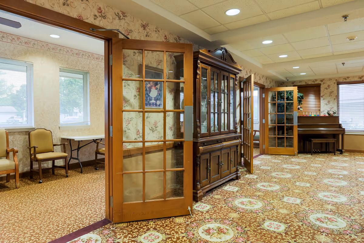 Interior view of a senior living facility room with floral patterned carpet and wallpaper. The room features wooden framed glass doors, a large wooden china cabinet, a piano with a bench, and several chairs near windows with blinds. The ceiling has recessed lighting.