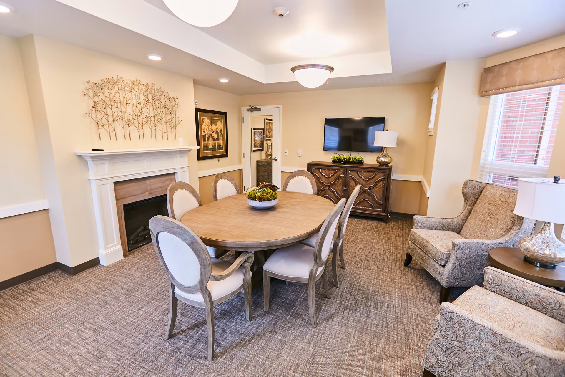 A cozy interior room featuring a round wooden table surrounded by six upholstered chairs. The room includes a fireplace with decorative metal wall art above it, a flat-screen TV mounted on the wall, a wooden cabinet beneath the TV, and two patterned armchairs with a side table and lamp near a window with blinds.