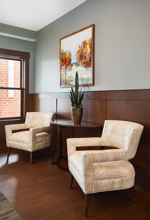 Two beige patterned armchairs positioned around a small round wooden table with a potted plant on it, set against a wall with wooden paneling and a large window showing a brick exterior. Above the table hangs a colorful abstract painting.