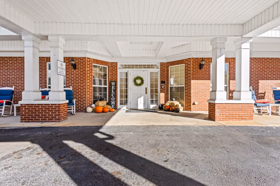 Entrance to a senior living facility with a covered porch supported by white columns. The building has red brick walls and a white door decorated with a green wreath. There are pumpkins and hay bales arranged on either side of the door, along with a vertical welcome sign. Blue cushioned chairs are placed along the porch area.