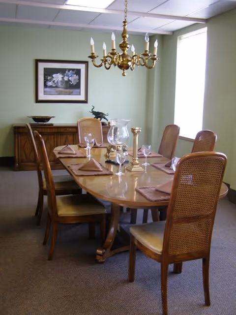 A dining room with a wooden table set for six people. The table has six wooden chairs with woven backs, each place setting includes a folded napkin and a wine glass. A brass chandelier hangs above the table. There is a sideboard against the wall with decorative items and a framed floral painting above it. A large window with white curtains allows natural light into the room.