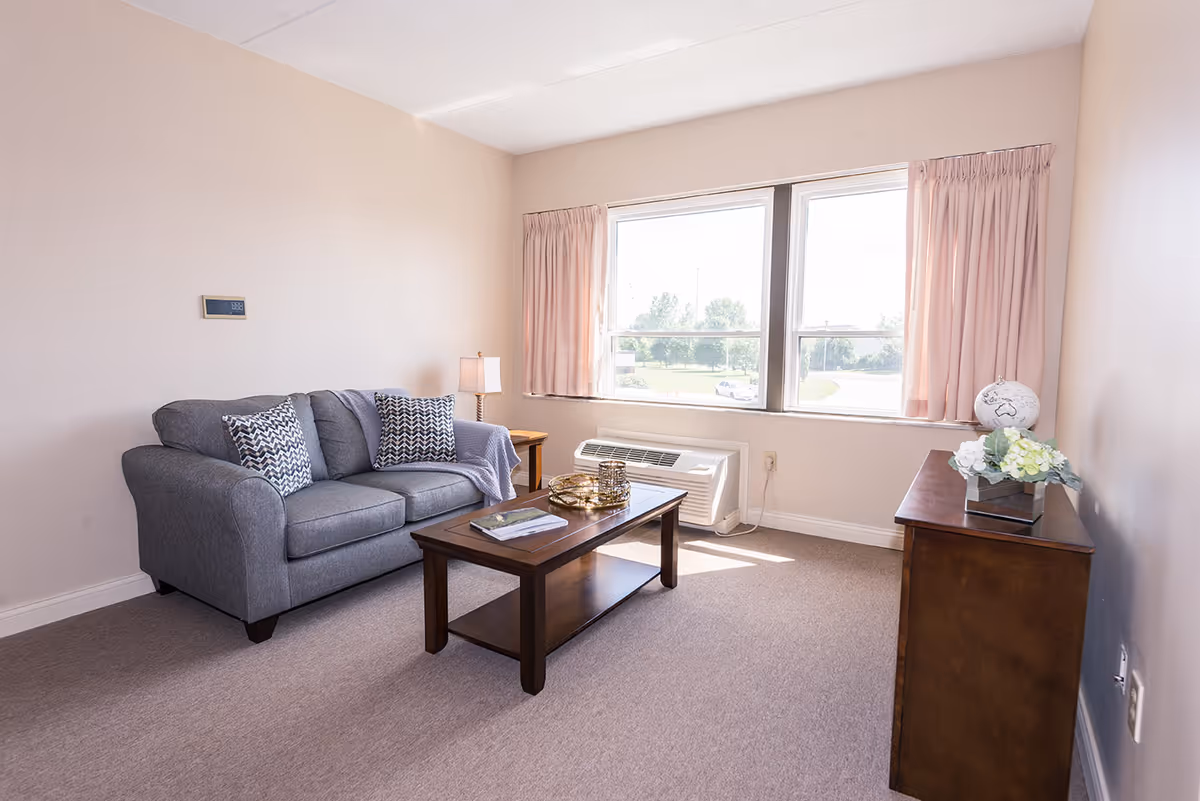 A bright living room with a gray couch adorned with patterned pillows, a wooden coffee table with decorative items, a side table with a lamp, a window with pink curtains letting in natural light, and a wooden cabinet with a globe and flower arrangement on top.
