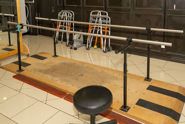 A rehabilitation exercise area with parallel bars mounted on a wooden platform, a black adjustable stool in front, and several walkers lined up against a dark paneled wall in the background.