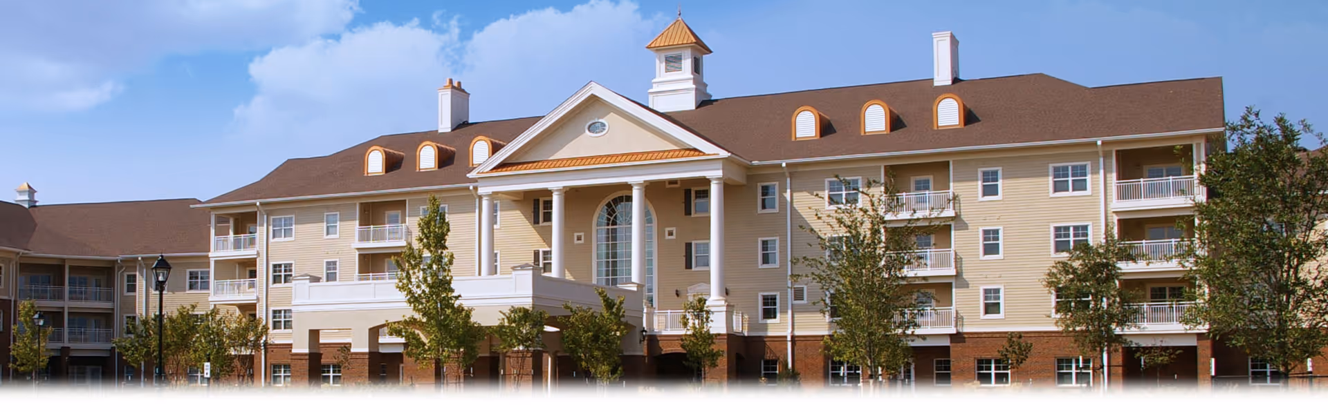 Exterior view of a large senior living facility building with beige siding, white columns, multiple balconies, and a brown roof under a partly cloudy sky.