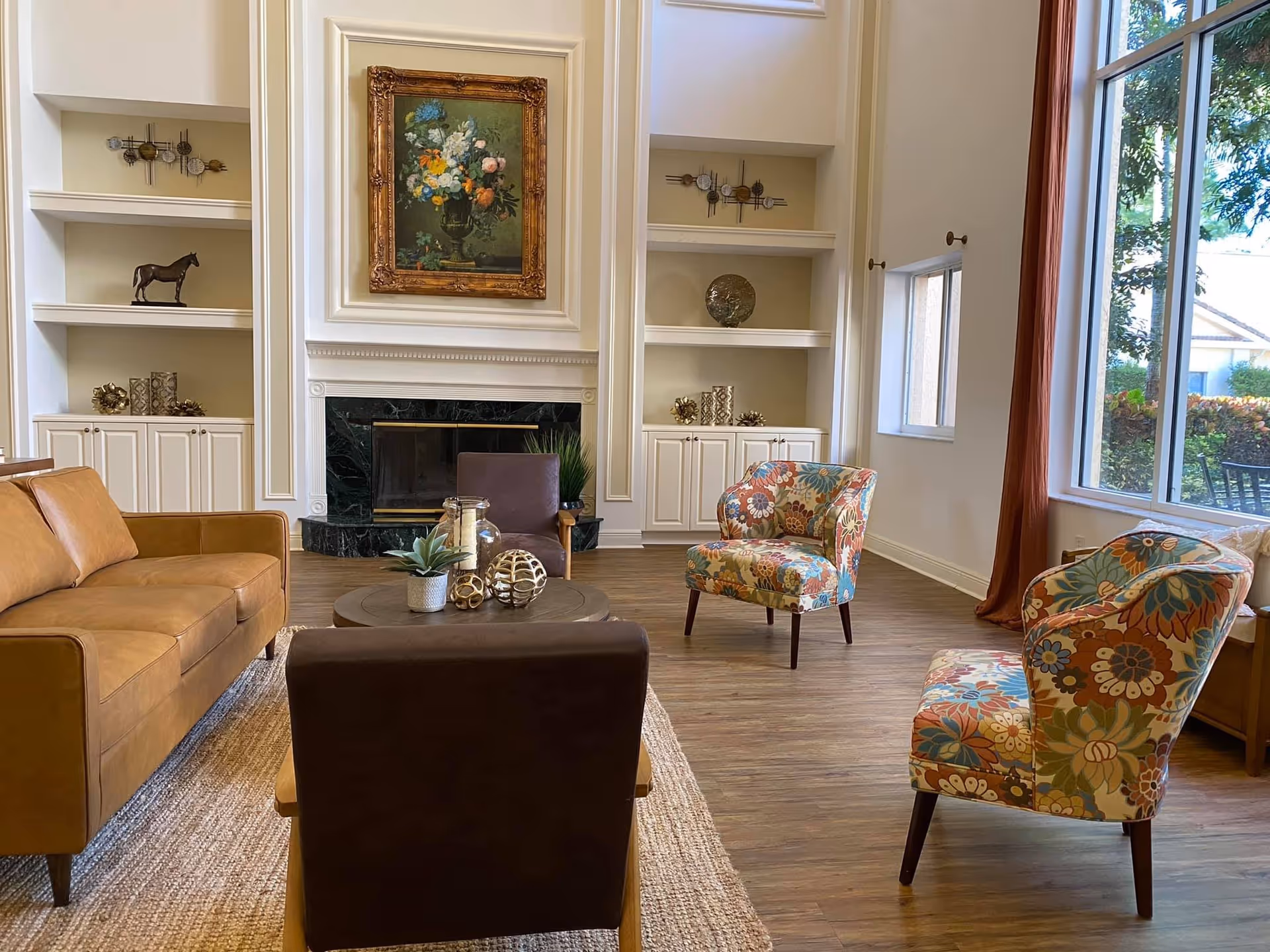 Bright furnished common living room with a leather sofa, patterned armchairs, coffee table, fireplace and large windows.