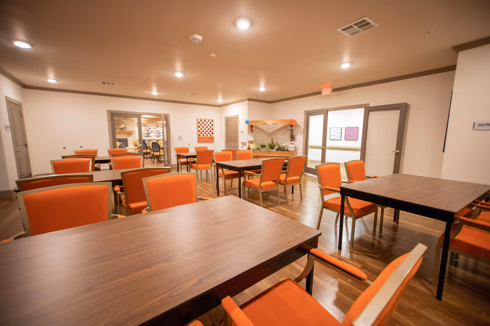 A spacious room with multiple wooden tables and orange cushioned chairs arranged neatly on a wooden floor. The room has beige walls with gray trim, recessed ceiling lights, and several doors and windows. There is a decorative plant display in the corner and framed artwork on the walls.