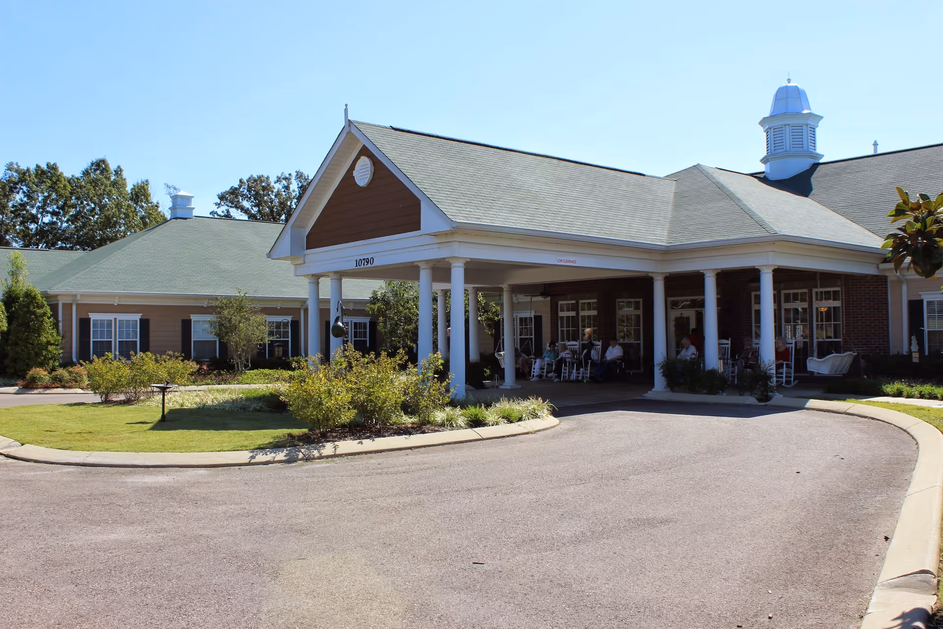 Front exterior view of SomerOak Senior Living facility showing a covered entrance with white columns, a circular driveway, and some people sitting on rocking chairs under the covered area. The building has a green roof, brick and siding walls, and is surrounded by landscaped bushes and trees.