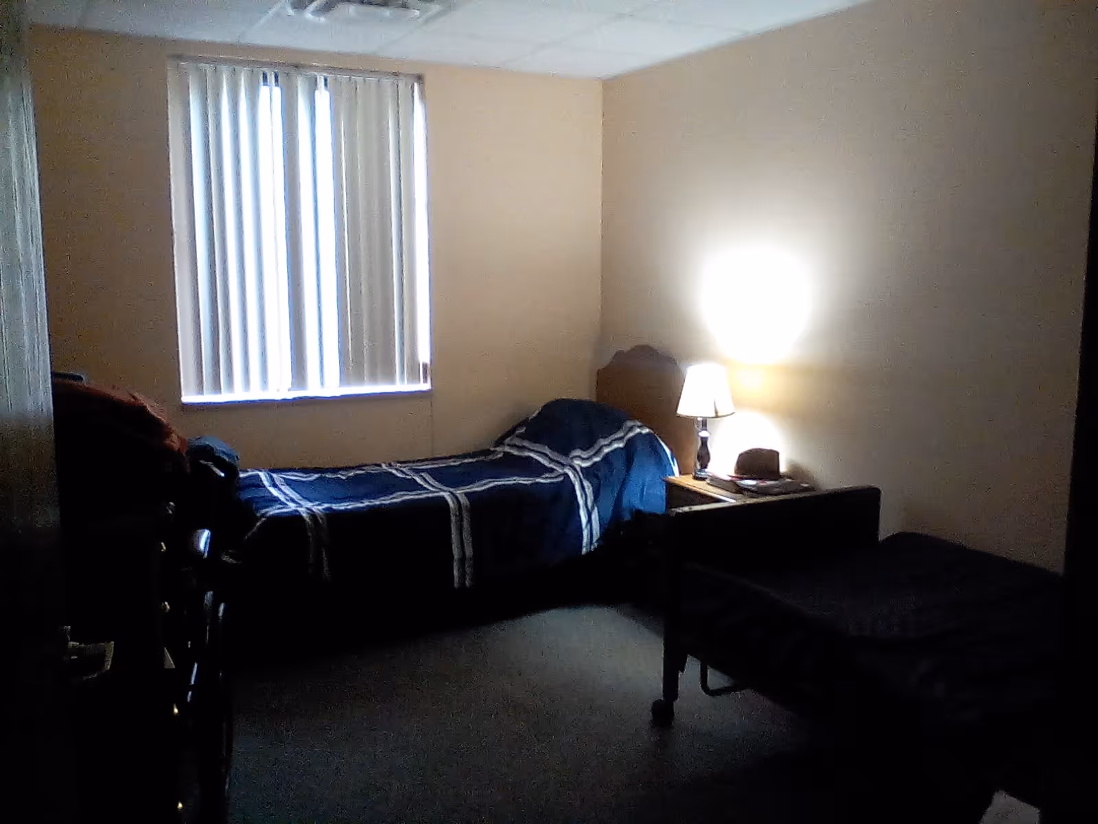 A dimly lit bedroom with two single beds. One bed has a blue and white striped blanket and is positioned near a window with vertical blinds. A small wooden nightstand with a lit lamp and a hat on it is placed between the beds. The walls are beige, and the floor is carpeted.