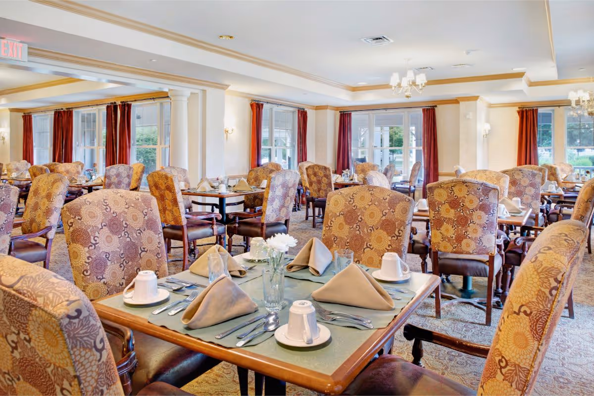 A bright and spacious dining room in a senior living facility with multiple tables set for meals. Each table has patterned upholstered chairs, beige napkins folded neatly, white cups, plates, and silverware. Large windows with red curtains allow natural light to fill the room, and chandeliers hang from the ceiling.