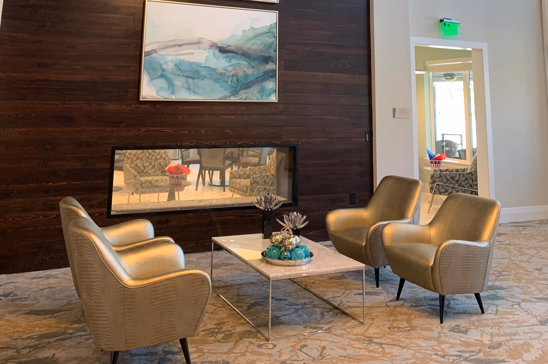 A cozy seating area with four modern armchairs arranged around a rectangular marble coffee table. The table has decorative items including a tray with blue and silver spheres and two silver flower sculptures. Behind the seating area is a dark wooden wall with a built-in horizontal fireplace and an abstract blue and white painting above it. To the right, there is a doorway leading to another room with additional seating and a green exit sign above the door.