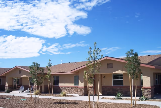 Single-story residential building with beige walls and brown roof under a blue sky with scattered clouds. Small trees and desert landscaping are in front of the building with a paved walkway.