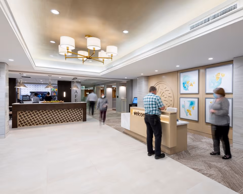Reception lobby with a welcome desk, café counter, wall artwork, and several people walking and standing.