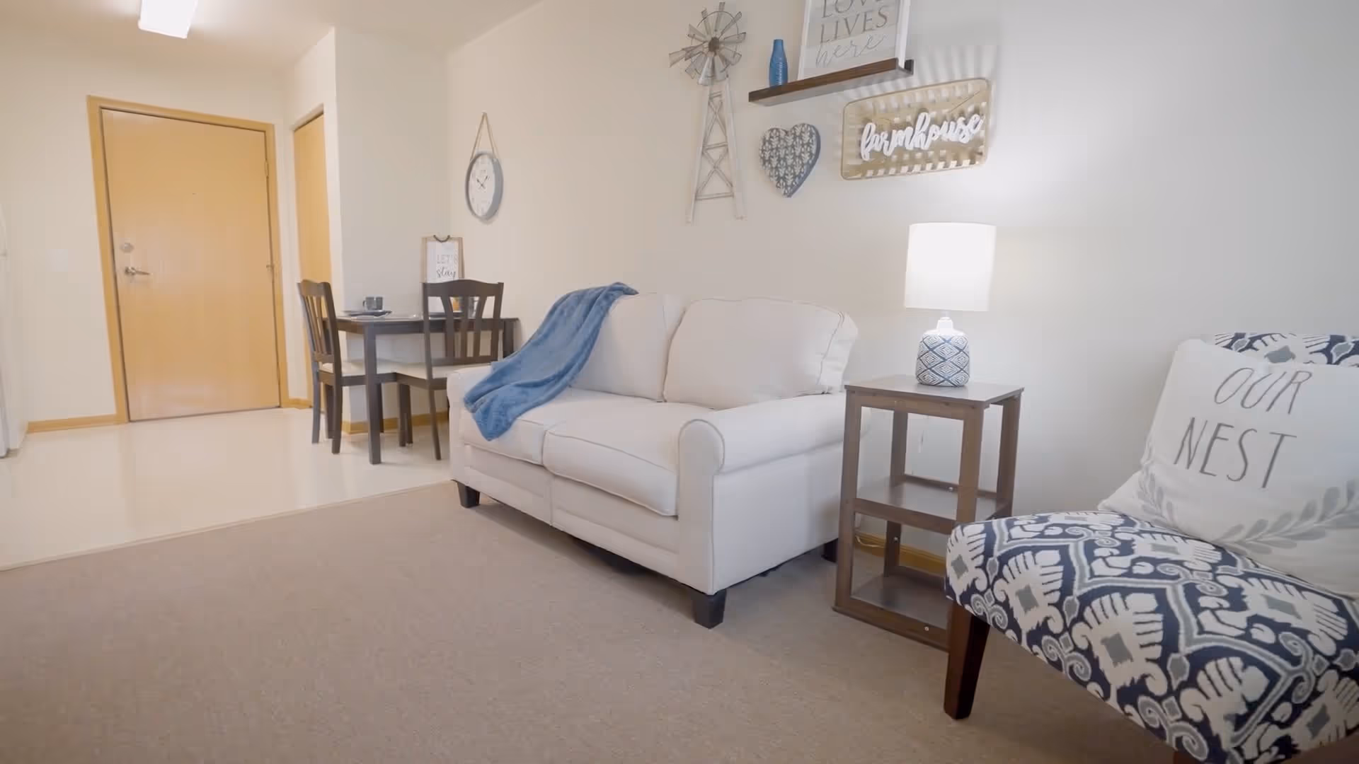 Bright living room with a white loveseat, patterned accent chair, side table with a lamp, and a small dining table near the entry door.