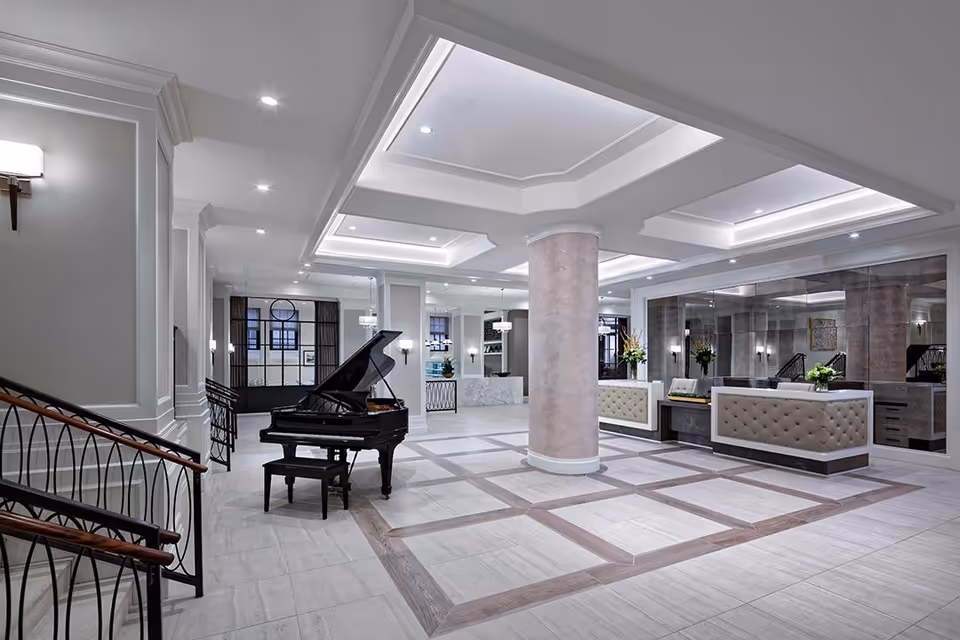 Spacious and elegant interior lobby area with a grand piano, a central column, modern recessed ceiling lights, and a reception desk with tufted upholstery. The space features large mirrors, light-colored tiled floors with wood accents, and a staircase with decorative railings.