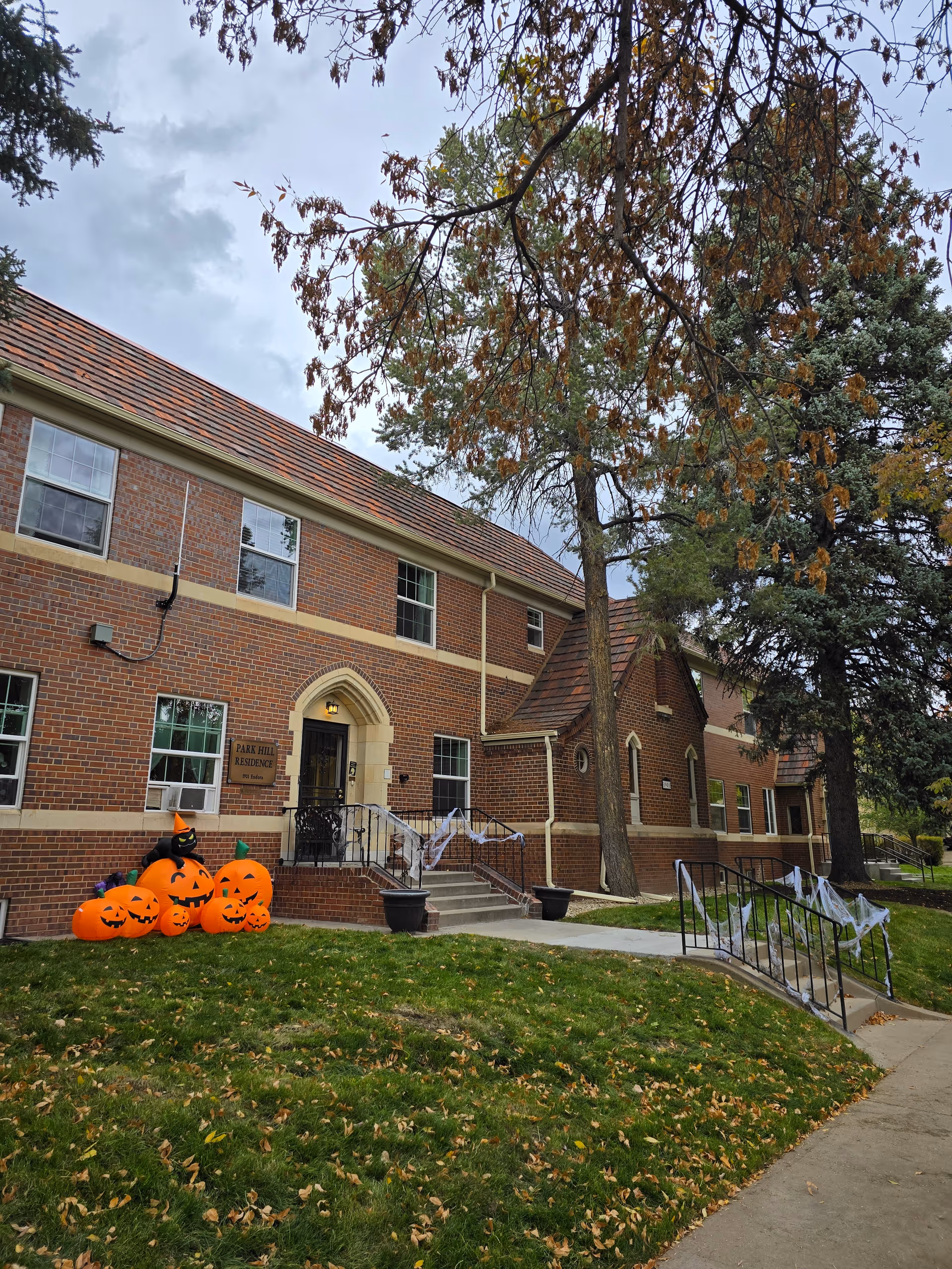 Exterior view of Park Hill Residence, a two-story brick building with a sloped roof. The entrance has steps and a ramp decorated with white spider webs for Halloween. Inflatable Halloween pumpkins and a black cat are displayed on the lawn near the entrance. Trees with autumn leaves surround the building under a cloudy sky.