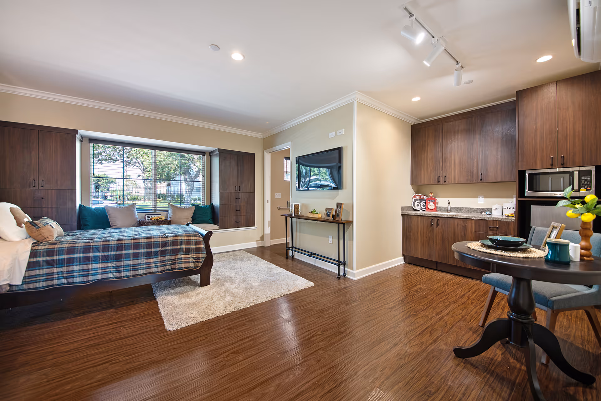 A cozy studio apartment interior with a single bed covered in a plaid blanket and pillows, a large window with built-in cabinets on either side, a small round dining table with two chairs, and a kitchenette with wooden cabinets, a microwave, and a mini fridge. The room has wooden flooring, a light-colored rug, and a wall-mounted TV above a narrow console table.