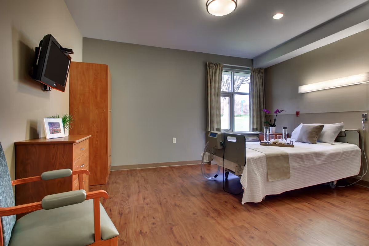 A clean and well-lit senior living bedroom with a hospital-style bed covered in white linens and beige accents. The room features a window with curtains, a wooden dresser with a framed picture and a small plant, a mounted flat-screen TV, and a green cushioned chair with wooden armrests. The floor is wood laminate, and there is a ceiling light fixture.
