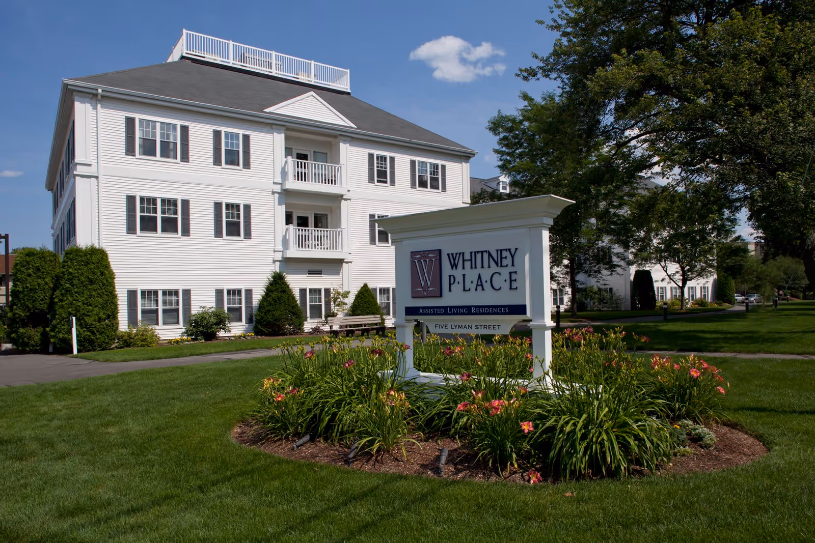 Exterior view of a white multi-story building with multiple windows and balconies, surrounded by green grass, trees, and flower beds. In the foreground, there is a white sign that reads 'Whitney Place Assisted Living Residences Five Lyman Street' under a clear blue sky.