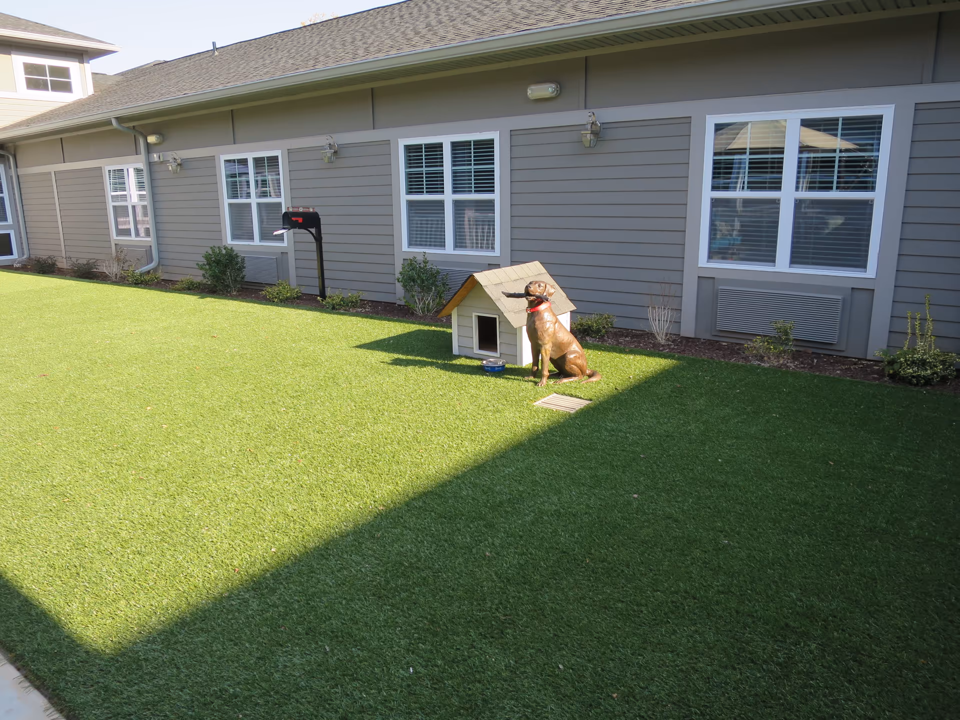 Outdoor area of Northlake House featuring a green lawn with a small doghouse and a statue of a dog wearing a red collar sitting next to it. The building exterior has gray siding with multiple windows and outdoor lights mounted on the wall.