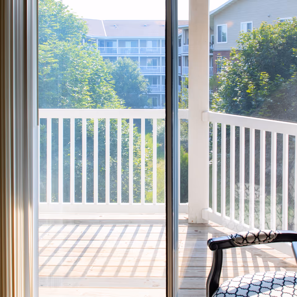 View through a sliding glass door onto a sunlit balcony with white railings and a patterned chair, overlooking trees and neighboring buildings.