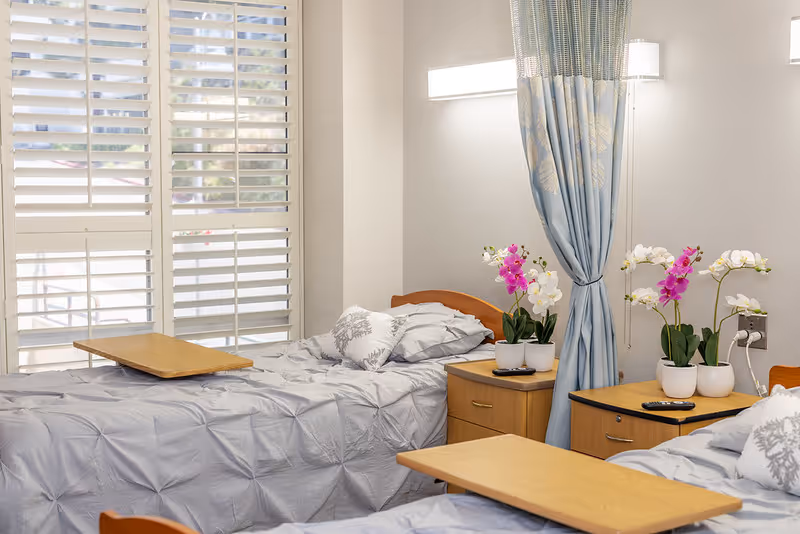 A bright room with two single beds covered in light gray bedding, each with a wooden overbed table. Between the beds are two wooden nightstands with potted orchids and remote controls. A blue curtain hangs between the beds, and large windows with white shutters let in natural light.