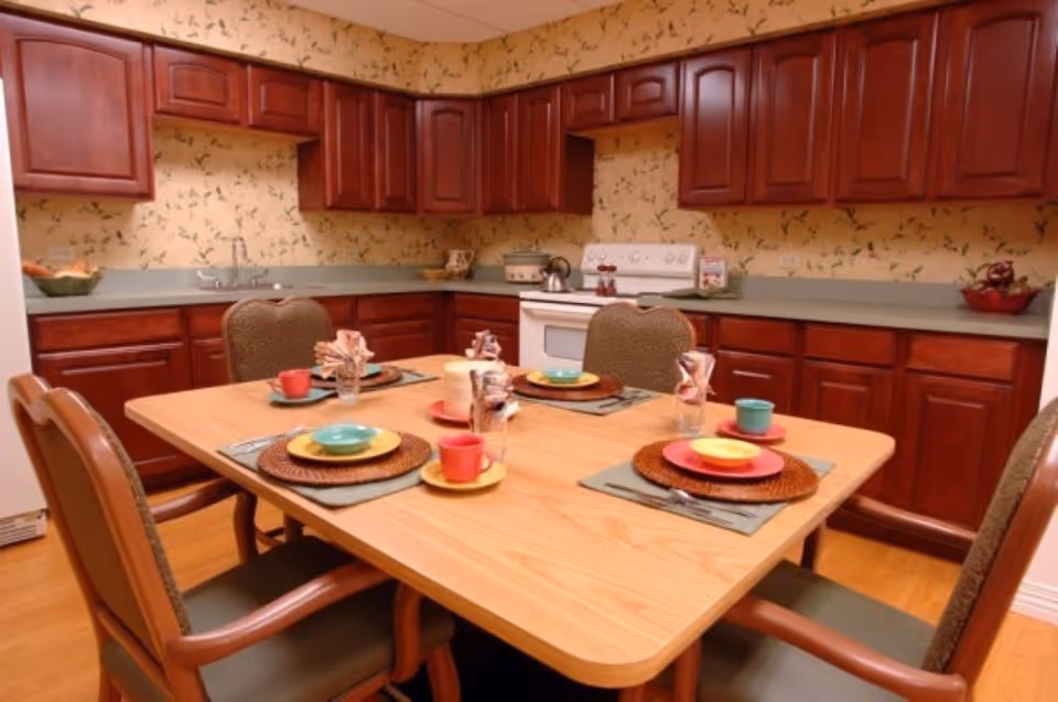 A kitchen and dining area with wooden cabinets and a light wood dining table set for four. The table has colorful plates, bowls, cups, and napkins arranged on placemats. The kitchen features a white stove, a double sink, and floral wallpaper.