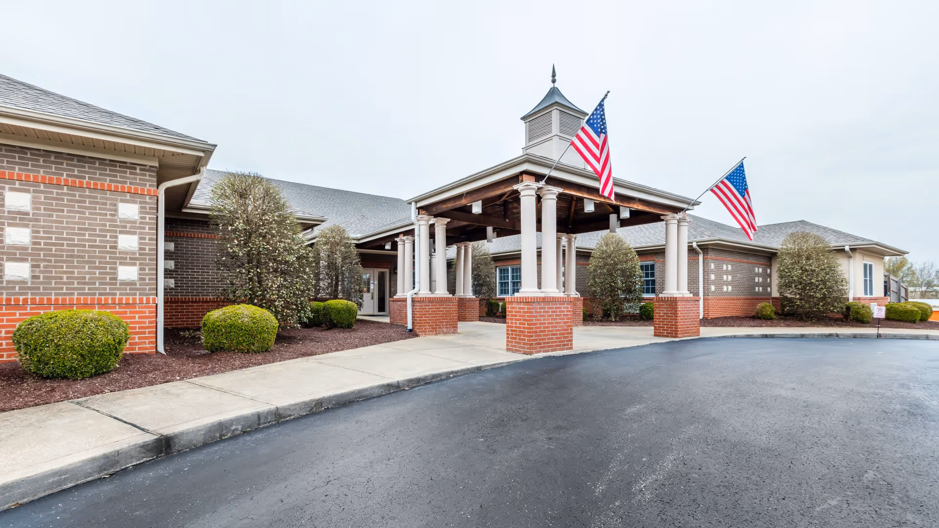 Exterior view of Wellington Manor Jackson showing the front entrance with a covered drop-off area supported by white columns on brick bases. Two American flags are mounted on the structure. The building is made of brick with neatly trimmed bushes and a paved driveway in front.