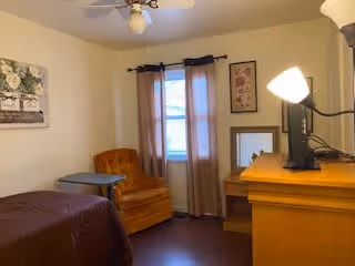 A small, cozy bedroom with a single bed covered in a brown blanket, an orange armchair next to a small round table, a window with beige curtains, a wooden dresser with a TV and a lamp on top, and framed artwork on the walls.