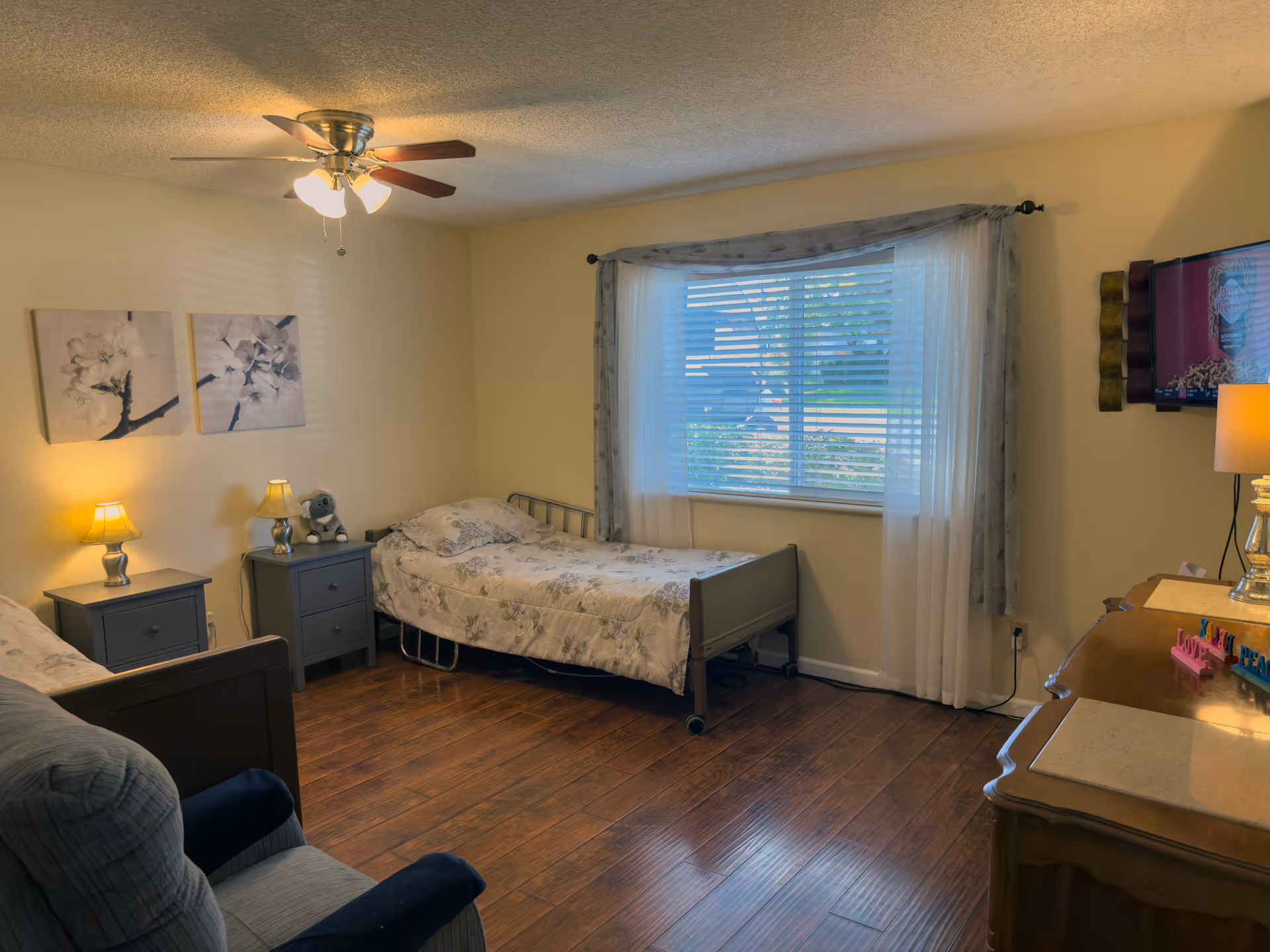 A cozy bedroom with two single beds, each with floral bedding. Between the beds are two matching nightstands with lamps and a small stuffed animal. There is a window with sheer curtains letting in natural light. A ceiling fan with lights is on the ceiling. A comfortable armchair is partially visible in the foreground, and a dresser with a lamp and a mounted TV is on the right side of the room. The floor is wooden.