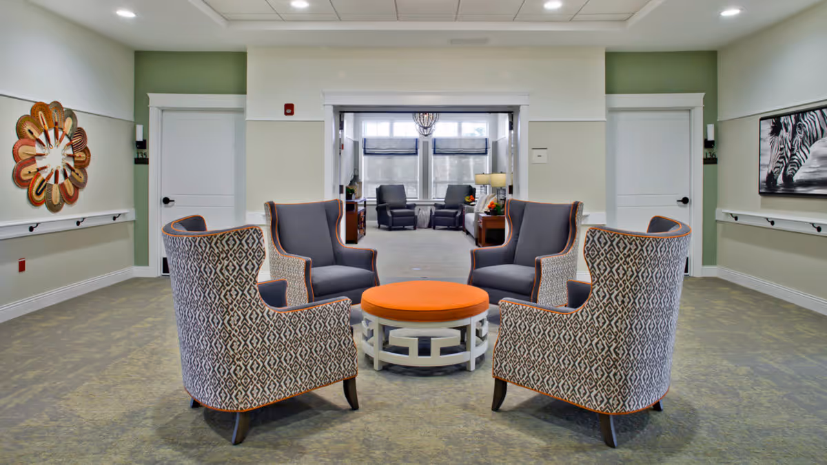 A seating area in a senior living facility with four patterned armchairs arranged around a round orange ottoman. The room has light green and white walls, two white doors, a decorative wall clock on the left, and a zebra painting on the right. In the background, there is another room with additional seating and large windows letting in natural light.