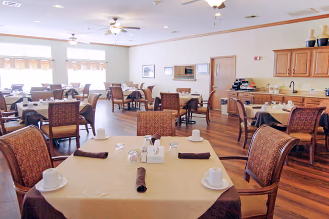 A bright dining room with multiple tables covered in beige and brown tablecloths, each set with cups, glasses, napkins, and condiments. The room has wooden flooring, large windows with valances, ceiling fans, and a kitchen area with wooden cabinets and appliances in the background.