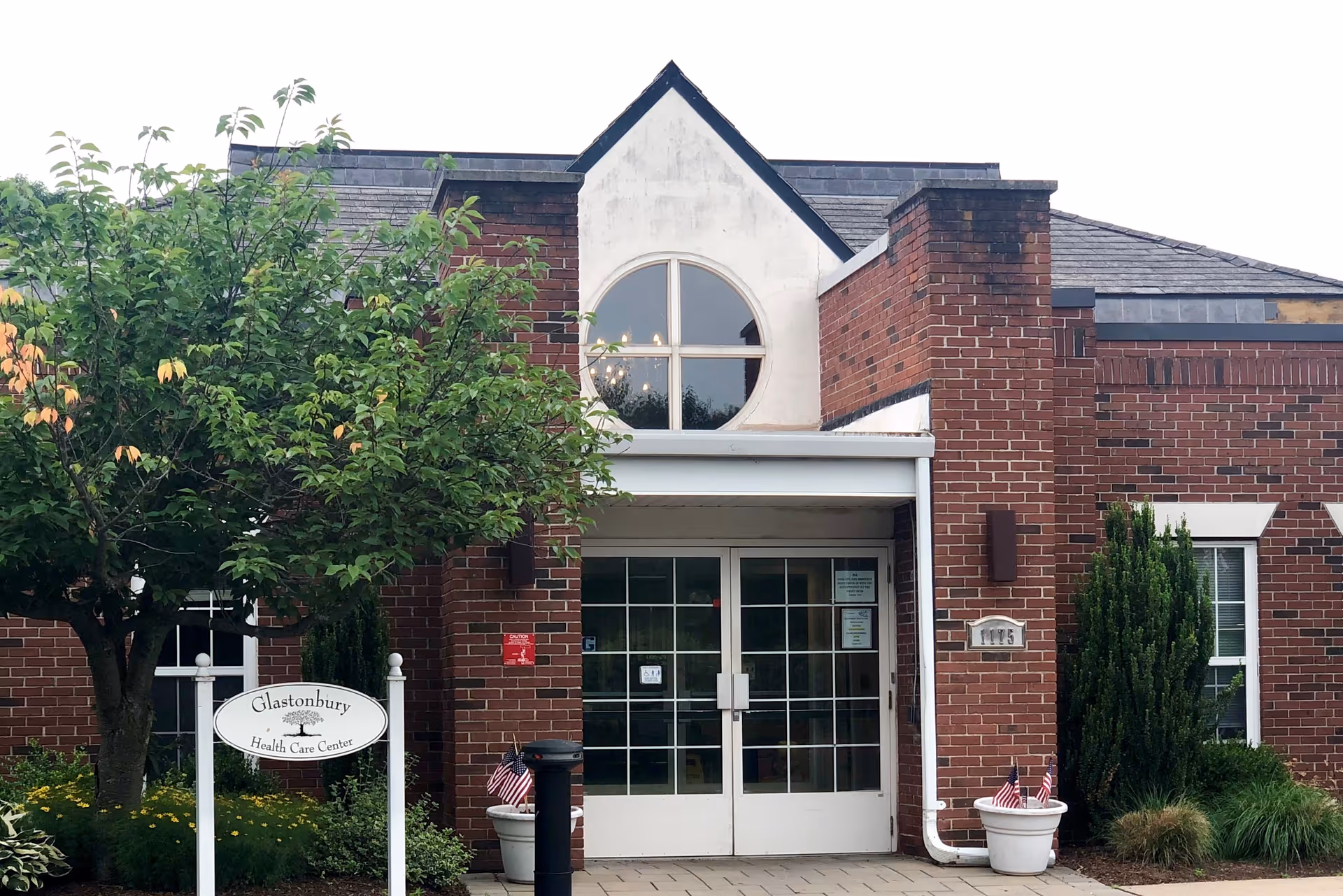 Front exterior view of the Glastonbury Health Care Center building with a brick facade, a white double-door entrance, a circular window above the entrance, and greenery including a tree and shrubs around the entrance. There is a white sign in front of the building that reads 'Glastonbury Health Care Center'.