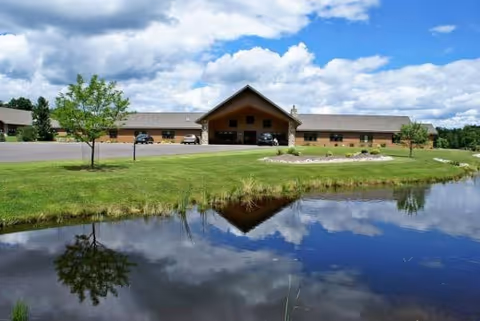 Front view of a single-story senior living facility with central entrance, parking, and reflective pond.