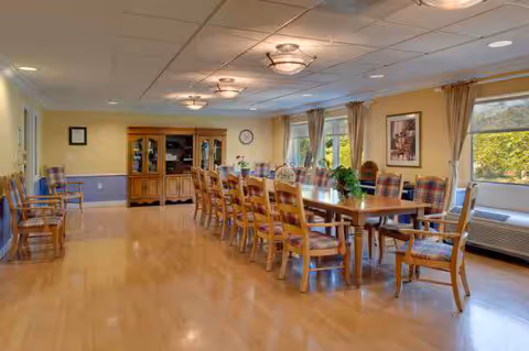 Communal dining room with a long wooden table surrounded by plaid-upholstered chairs, windows, and a buffet cabinet.