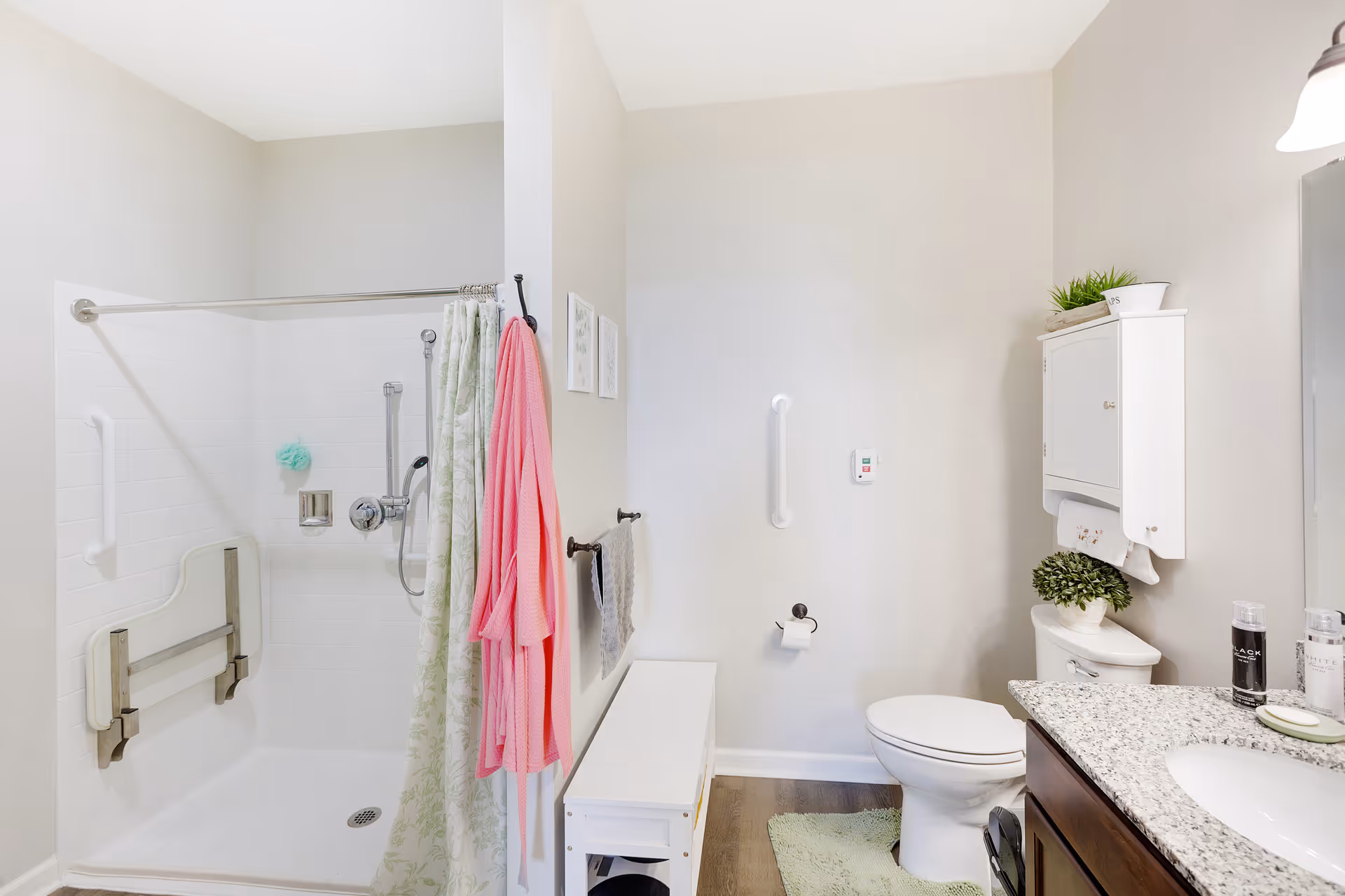 A clean and bright bathroom featuring a walk-in shower with a fold-down seat and grab bars, a shower curtain, a pink bathrobe hanging on the wall, a toilet with a small green plant on top, a white cabinet mounted on the wall, and a granite countertop with a sink and toiletries.