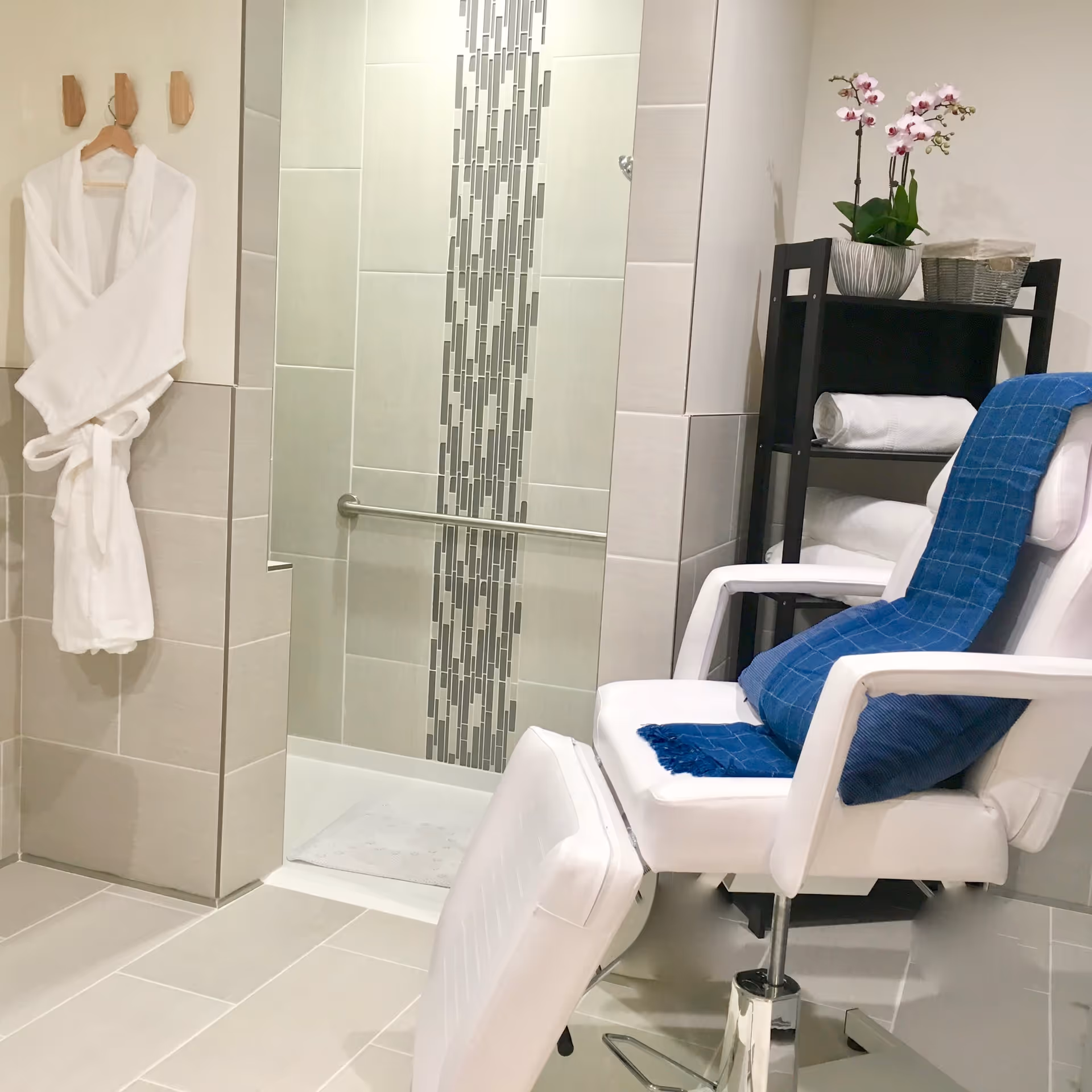 A clean and modern bathroom area featuring a walk-in shower with decorative vertical tile accents and a grab bar. A white bathrobe hangs on wooden hooks on the wall. In the foreground, there is a white reclining chair with a blue towel draped over it. A black shelving unit holds rolled white towels, a basket, and a potted orchid with pink flowers.