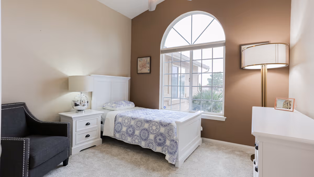 A cozy bedroom with a single white bed covered with a blue and white patterned quilt. Next to the bed is a white nightstand with a silver lamp. There is a black armchair with nailhead trim on the left side. The room has beige and brown walls, a large arched window letting in natural light, a floor lamp, and a white dresser with a small framed picture on top.