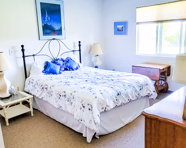 A bright bedroom with a double bed featuring a black metal headboard, white and floral bedding, and two blue decorative pillows. There are two bedside tables with lamps on either side of the bed, a wooden dresser, a window with a beige blind, and framed artwork on the walls.