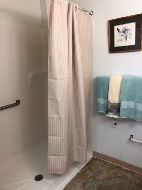 A bathroom with a beige shower curtain partially closed around a white shower stall. A metal grab bar is mounted inside the shower. On the wall outside the shower, there is a towel rack holding three folded towels in teal, cream, and patterned colors. Above the towel rack, a framed floral artwork is hung. The floor has a patterned mat near the shower entrance.