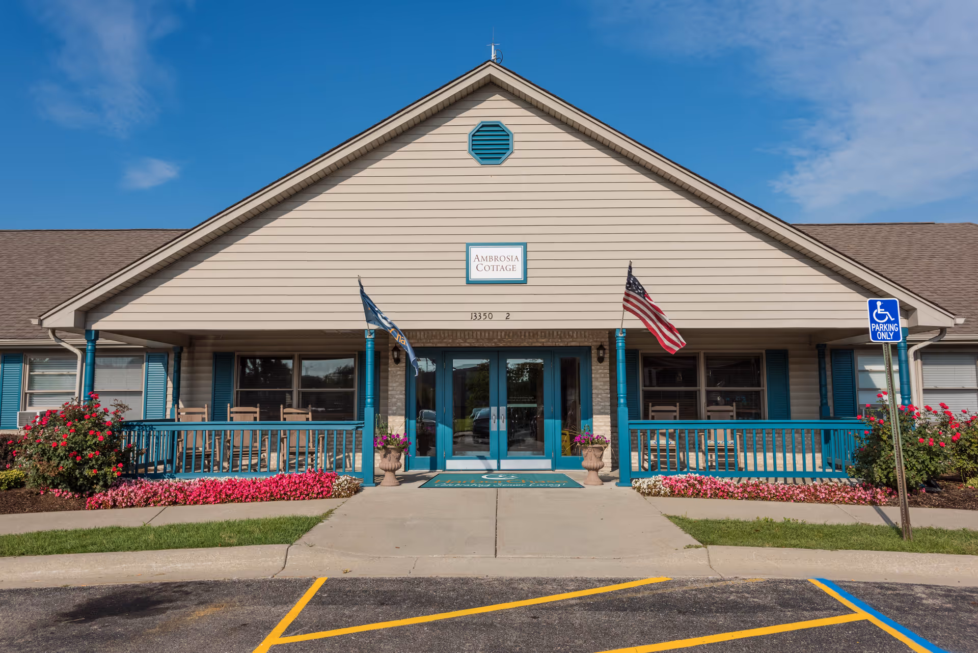 Front entrance of a single-story senior living cottage with a covered porch, teal railings, an American flag, and flower beds.