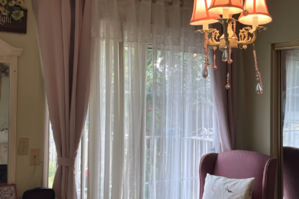 Interior living room with sheer curtains, pink chair, cushion, and ornate chandelier.