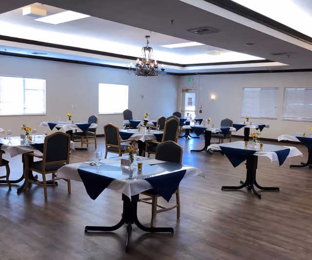 Spacious dining room with multiple small tables set with white and navy tablecloths, chairs, floral centerpieces, and a central chandelier.