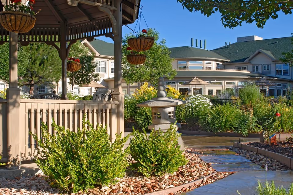 Outdoor garden area at Gardens at Columbine featuring a wooden gazebo with hanging flower baskets, lush green bushes, a stone lantern sculpture, and a paved walkway surrounded by various plants and flowers. The background shows a large building with green roofs and multiple windows under a clear blue sky.